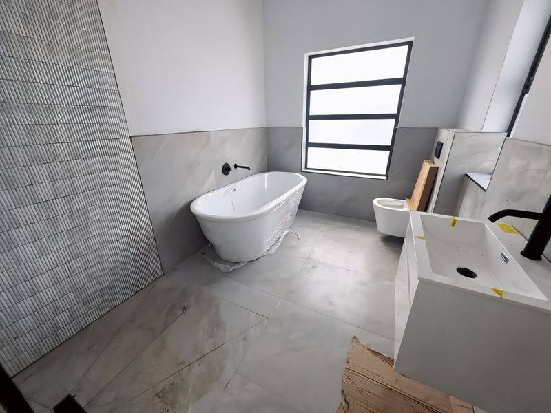 A modern, newly installed bathroom featuring a freestanding bathtub, wall-mounted faucet, vanity basin, and wall-mounted toilet with contemporary grey tiling.