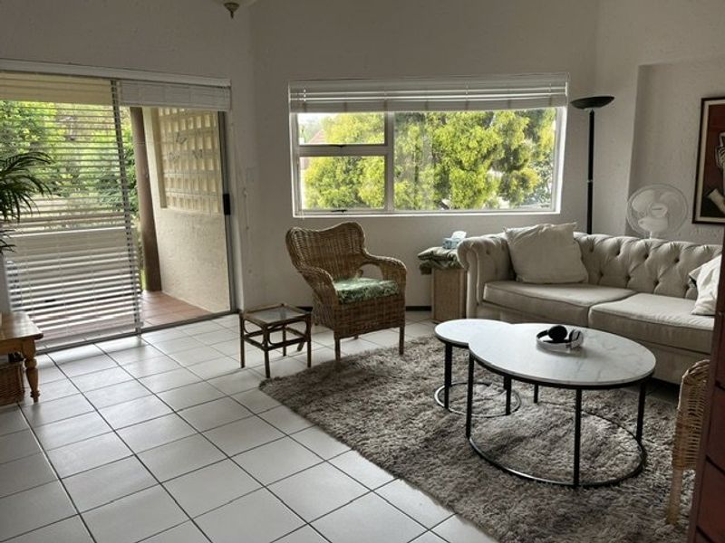 A bright living room featuring tiled flooring, large windows with venetian blinds, and a sliding glass door providing access to an outdoor patio area.