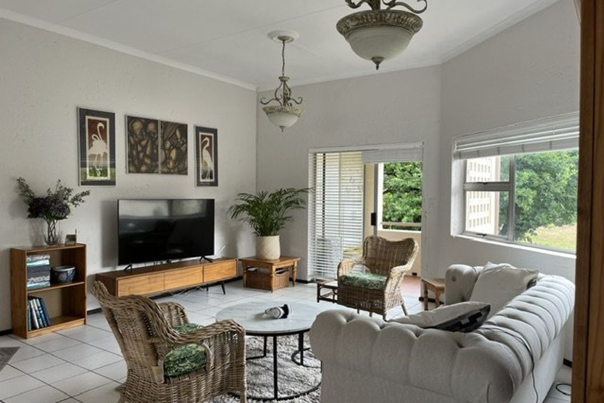 A bright living room featuring tiled flooring, large windows with venetian blinds, and direct access to an outdoor patio area.
