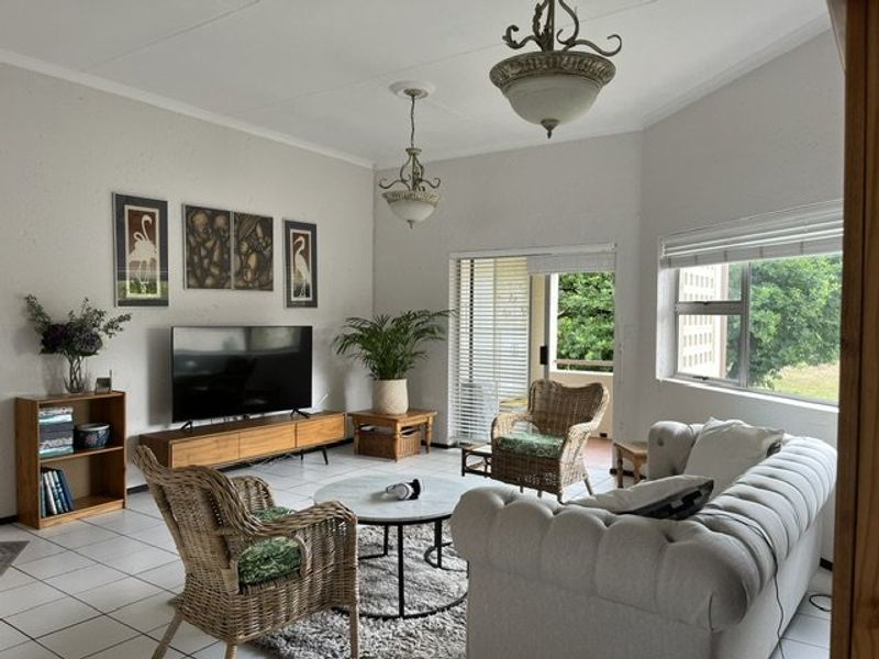 A bright living room featuring tiled flooring, large windows with venetian blinds, and direct access to an outdoor patio area.
