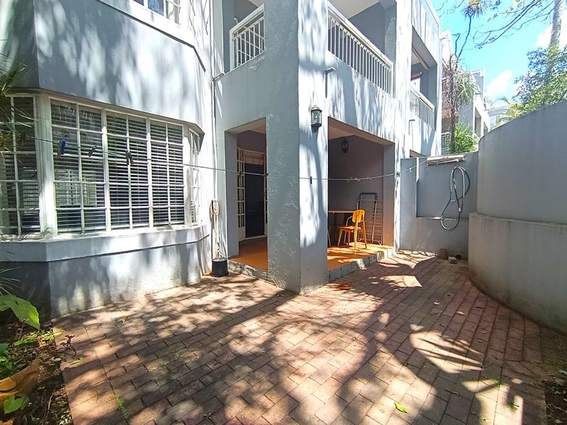 A rear exterior view of an apartment featuring a paved patio area, a covered balcony above, and exterior wall lighting.