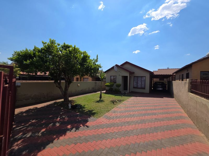 Front view of a residential property featuring a paved driveway, a carport, and a well-maintained garden area enclosed by a boundary wall.