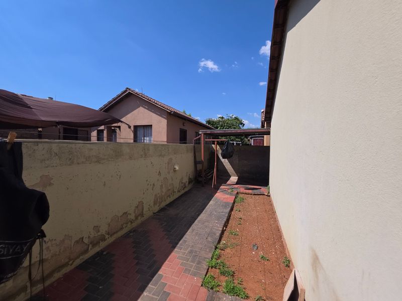 A narrow rear exterior side passage featuring a paved walkway, boundary wall with visible wear, and an adjacent soil area.