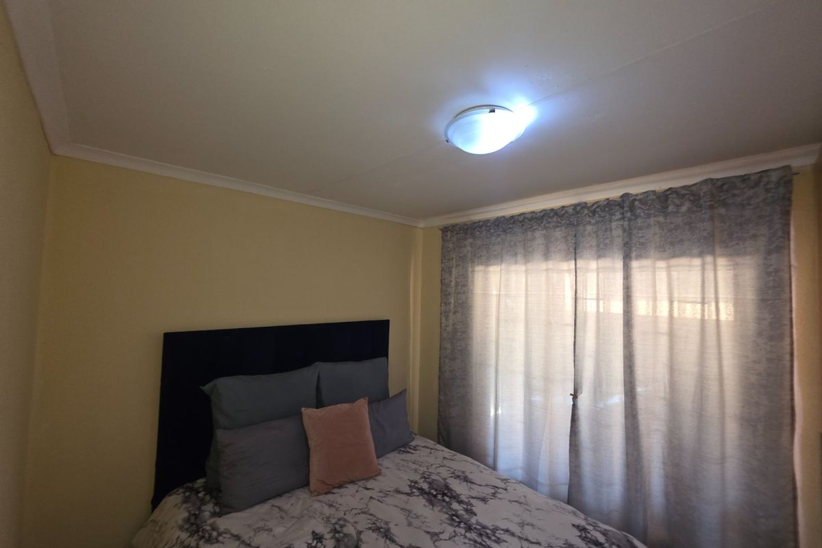 A standard bedroom featuring neutral wall finishes, ceiling cornicing, and a central flush mount light fixture with natural light filtering through window curtains.