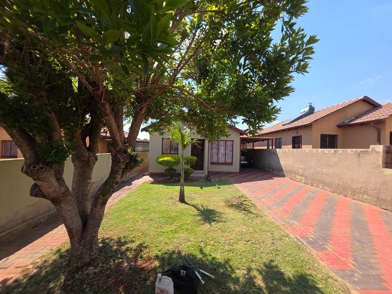 Front view of the property featuring a paved driveway, established garden, boundary wall, and a carport.