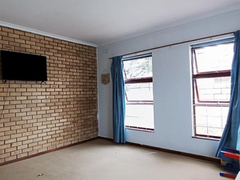 Living room featuring an exposed brick accent wall, large top-hung windows with security bars, and neutral carpeting.