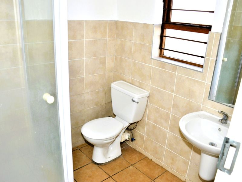 A functional bathroom featuring a shower enclosure, pedestal basin, and toilet with neutral ceramic wall and floor tiling.