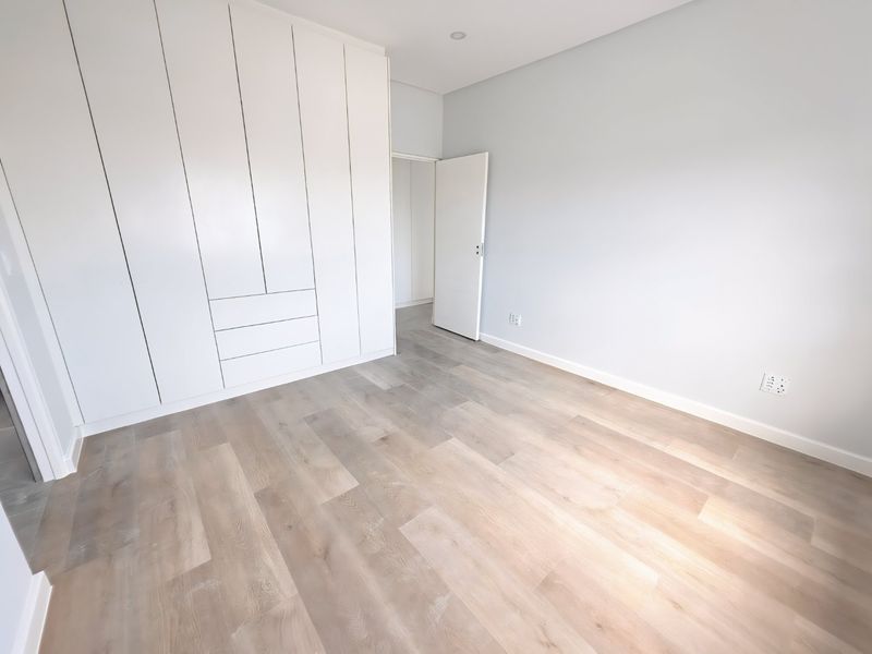 A bright, modern bedroom featuring light-toned laminate flooring, neutral walls, and extensive floor-to-ceiling built-in cupboards.