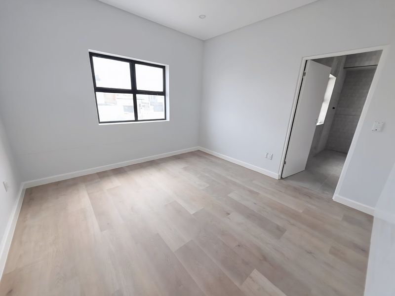 A bright, modern, and empty bedroom featuring light-toned laminate flooring, neutral walls, and an adjoining en-suite bathroom.