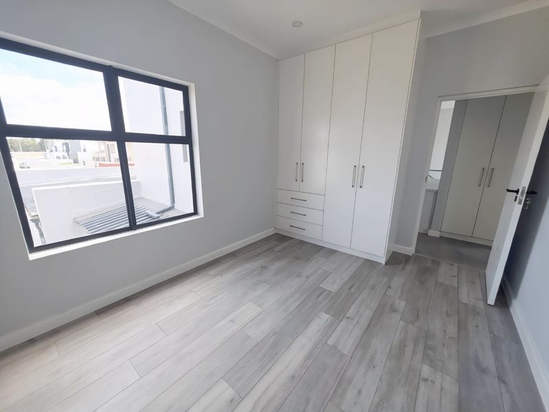 A bright, modern bedroom featuring light grey laminate flooring, built-in white cupboards, and large aluminium-framed windows.