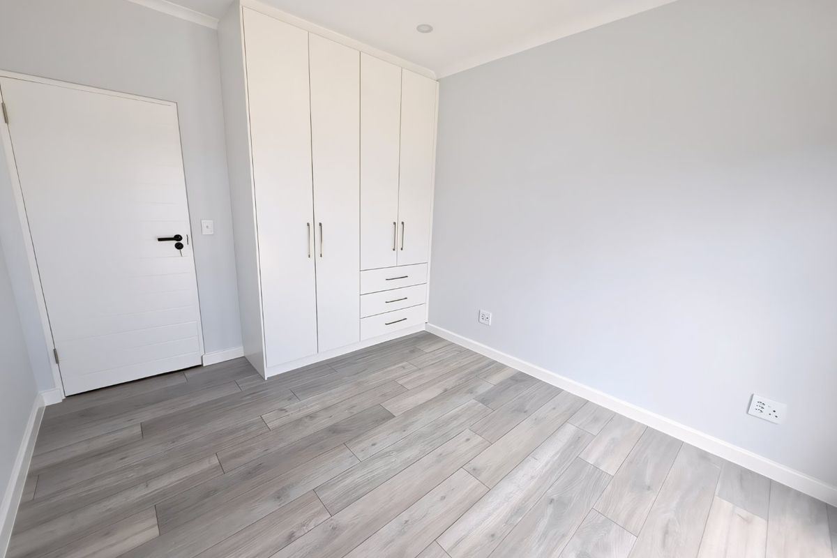 A bright, empty bedroom featuring modern light-toned laminate flooring, crisp white walls, and ample built-in cupboard storage.