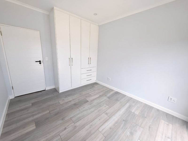A bright, empty bedroom featuring modern light-toned laminate flooring, crisp white walls, and ample built-in cupboard storage.