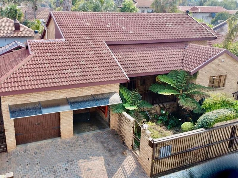 Aerial view of a brick townhouse featuring a tiled roof, paved driveway, garage, and a gated entrance with established garden landscaping.