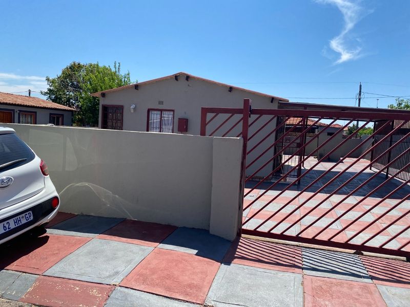 Front exterior view of the property featuring a paved driveway, secure metal gate, perimeter wall, and a carport structure.