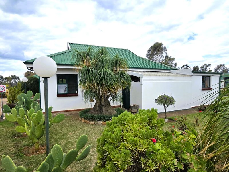Front view of a townhouse featuring a green metal roof, white masonry exterior, and a landscaped garden area with a paved driveway.