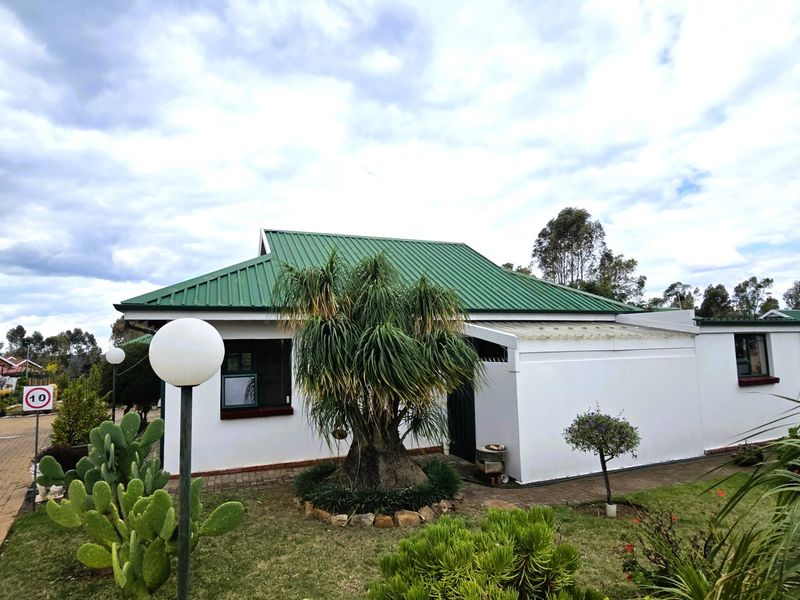 Front view of a townhouse featuring a green metal roof, paved driveway, and established garden area.
