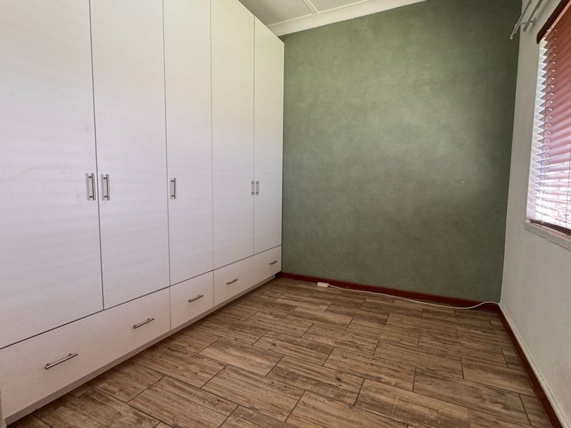A clean, empty bedroom featuring wood-look tiled flooring, a large white built-in wardrobe, and a green feature wall with venetian blinds.