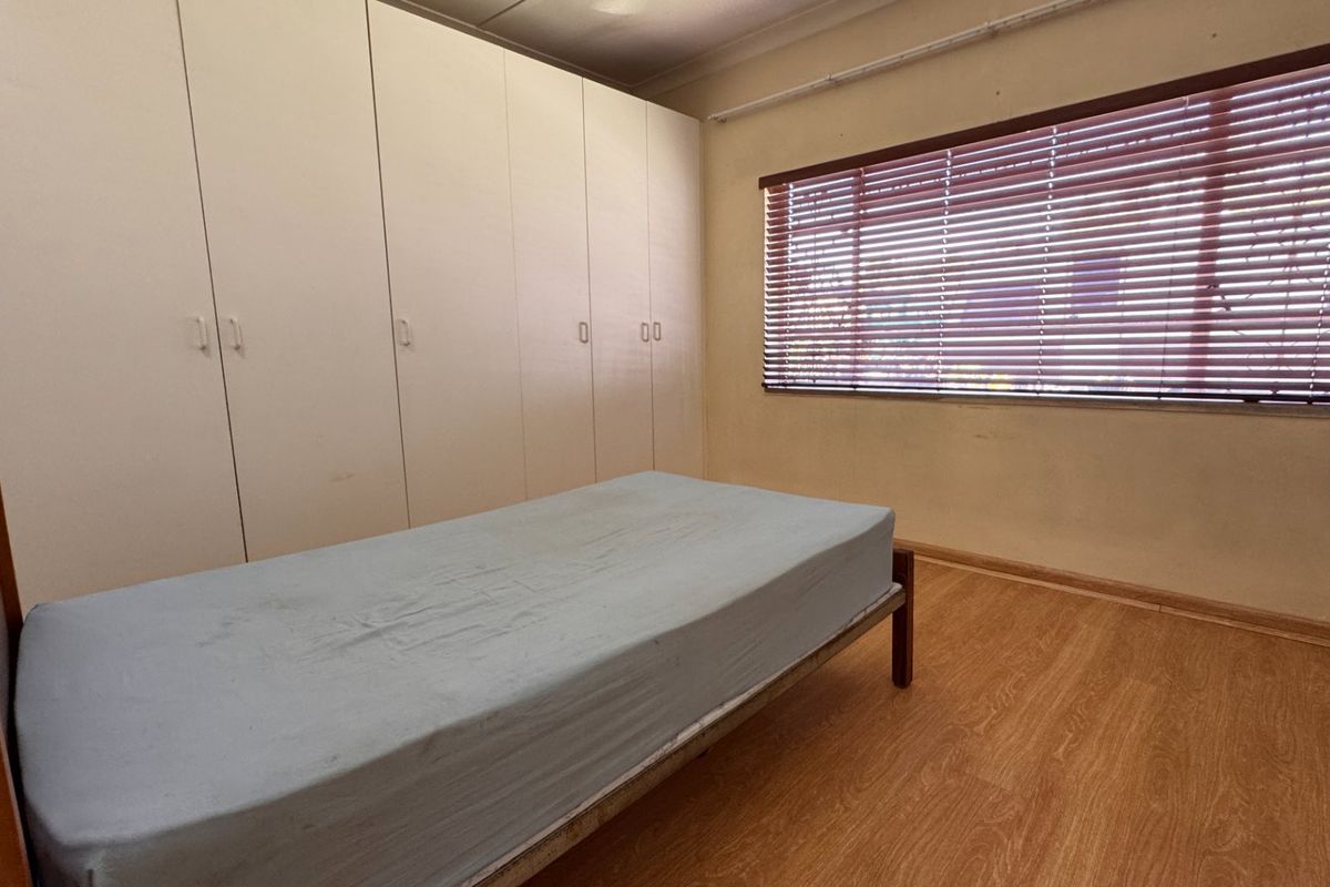 A bedroom featuring laminate flooring, built-in cupboards, and venetian blinds. The room offers a functional layout with neutral finishes.