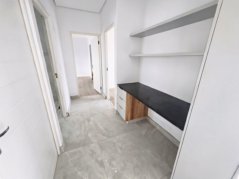 A bright, modern hallway featuring a built-in desk area with shelving and neutral tiled flooring, providing a functional workspace transition between rooms.