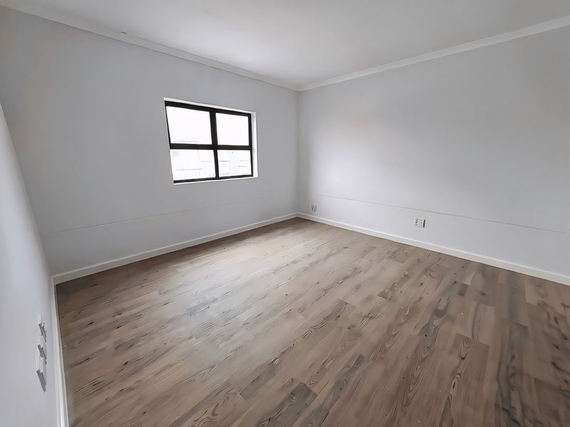 A bright, empty bedroom featuring modern laminate wood flooring, neutral walls, and a clean aluminium-framed window.