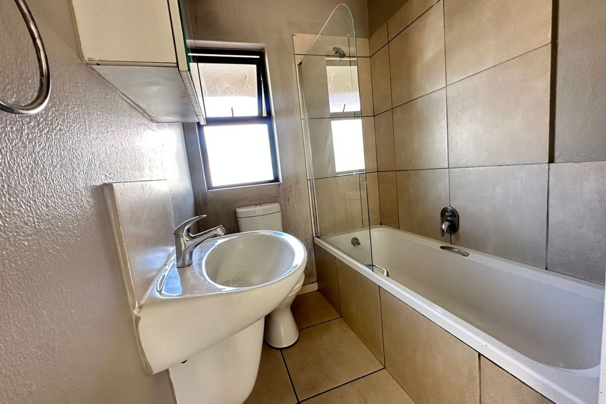 A functional bathroom featuring a pedestal basin, toilet, and a bathtub with a glass shower screen, finished with neutral wall and floor tiling.