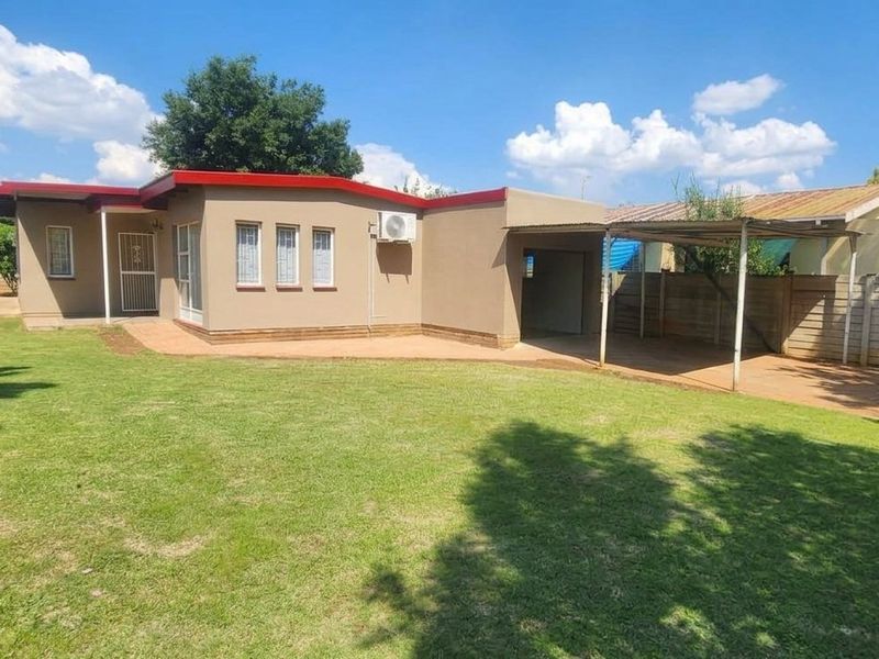 Rear view of the property featuring a spacious lawn, paved driveway, and a covered carport area with a boundary wall.