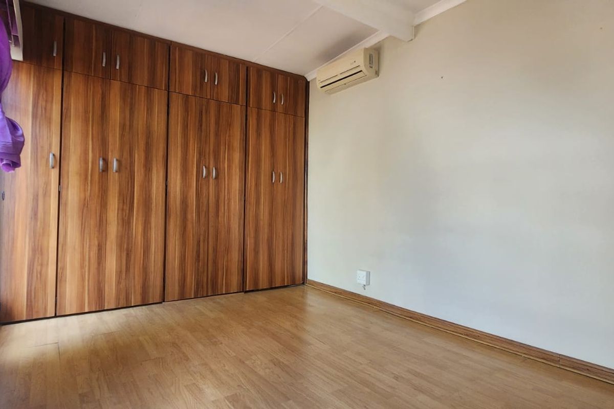 A bright, empty bedroom featuring wood-look laminate flooring, extensive built-in wooden cupboards, and a wall-mounted air conditioning unit.