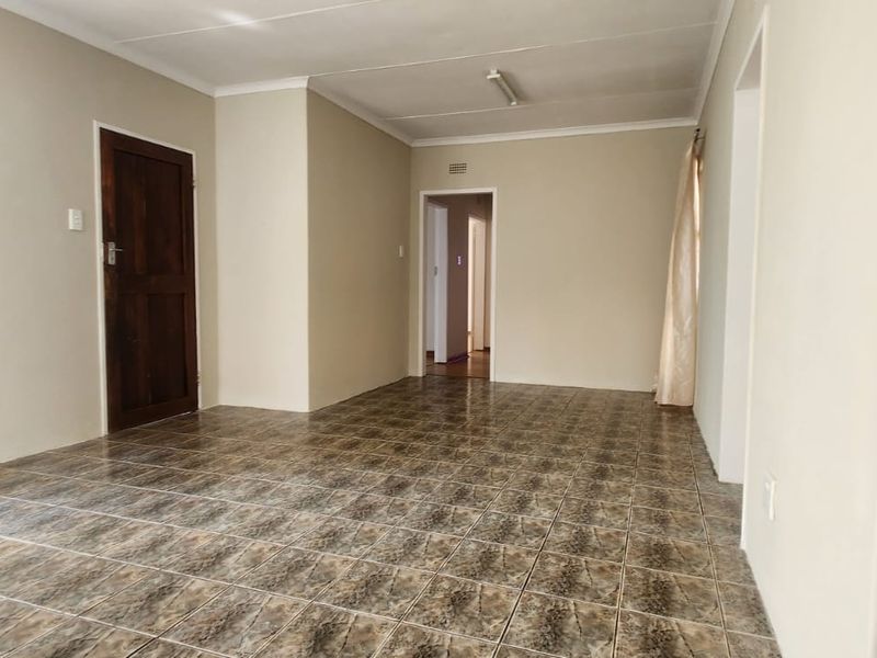 An empty living area featuring neutral wall paint, tiled flooring, and a wooden door leading to an adjacent room.