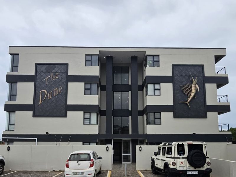 Front elevation of The Dune apartment complex featuring a paved parking area, modern facade, and secure perimeter wall with electric fencing.