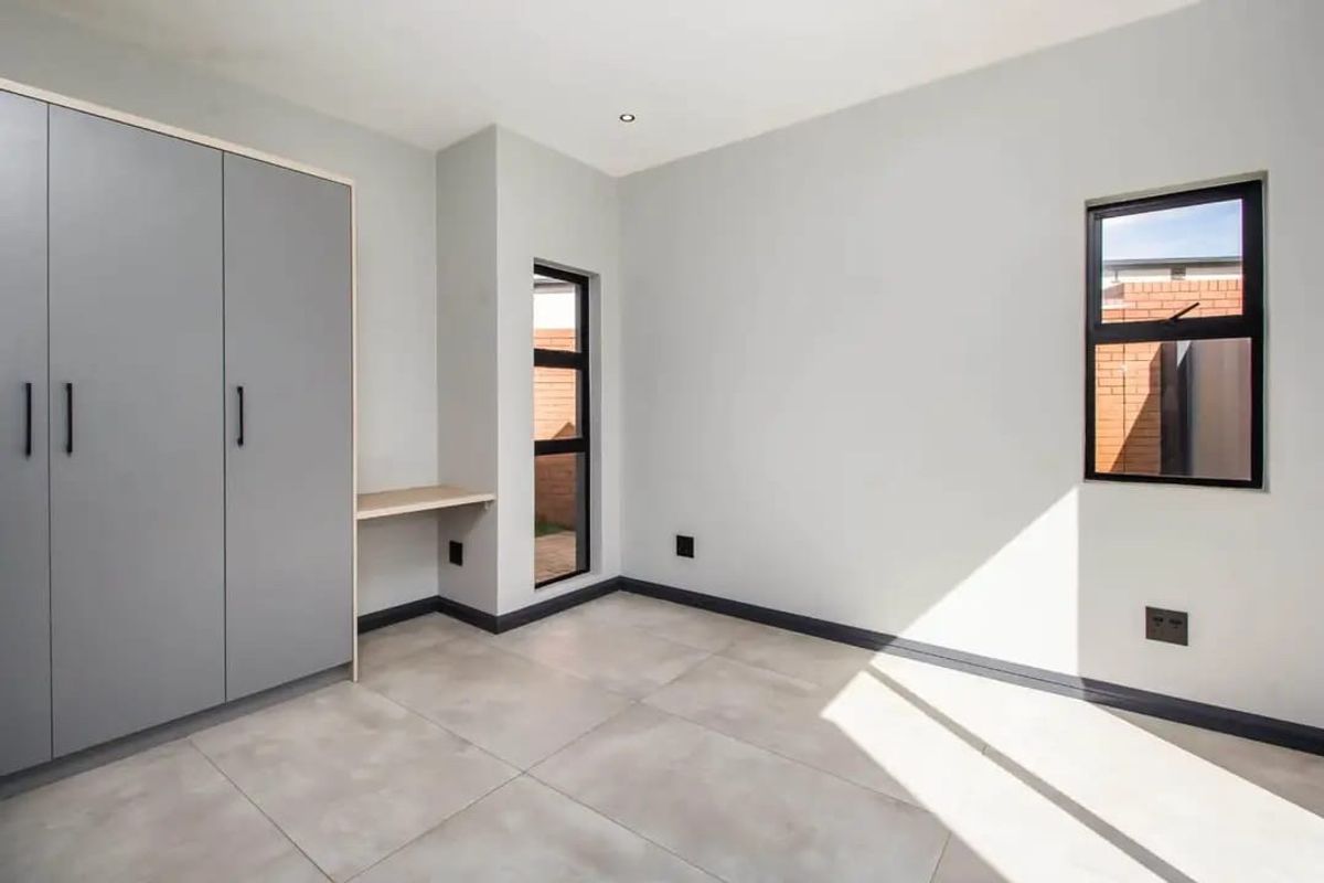 A bright, modern bedroom featuring neutral tiled flooring, built-in cupboards, a integrated desk area, and large aluminium-framed windows.