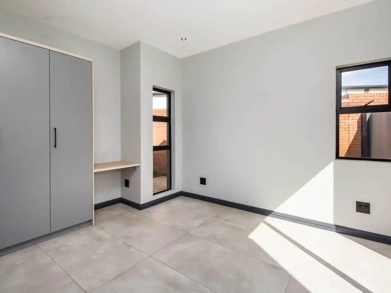 A bright, modern bedroom featuring neutral tiled flooring, built-in cupboards, a integrated desk area, and large aluminium-framed windows.