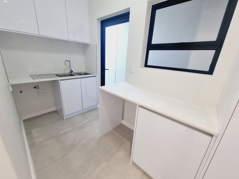 A modern, clean scullery featuring white built-in cabinetry, stone countertops, a stainless steel sink, and neutral tiled flooring with direct outdoor access.