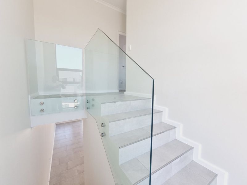 A modern, minimalist staircase featuring light grey floor tiles and a sleek frameless glass balustrade with stainless steel fixings.