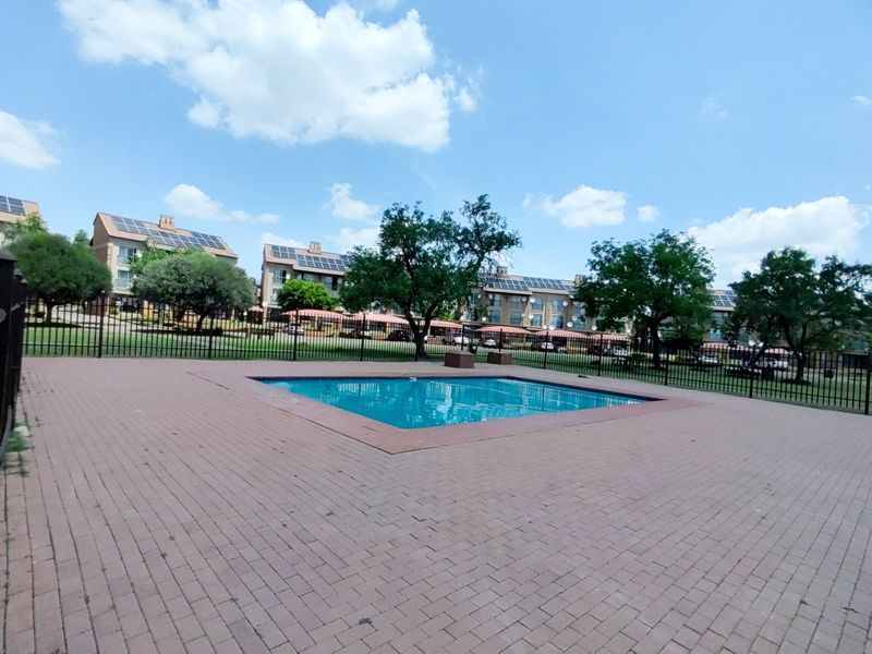 A communal swimming pool area featuring a paved deck, perimeter fencing, and views of the surrounding apartment complex with solar-equipped roofs.