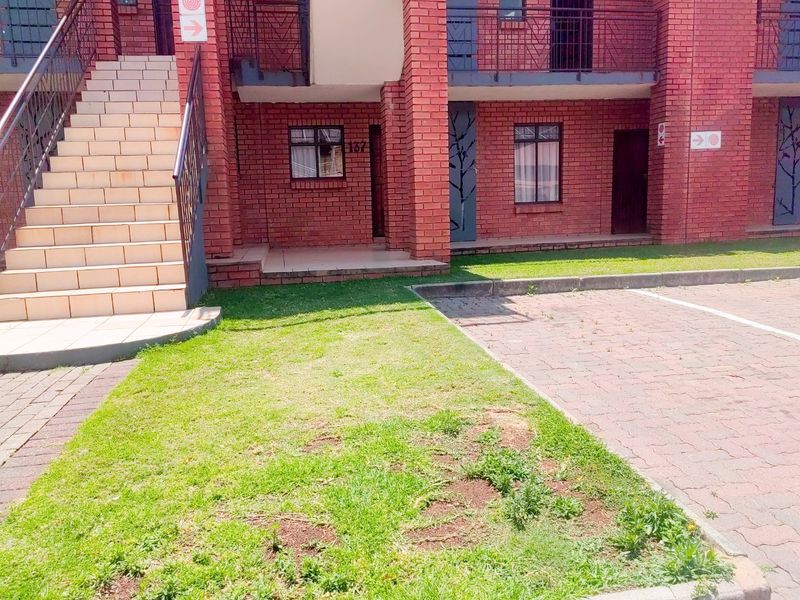 Exterior view of a face brick apartment building featuring a tiled staircase, metal balustrades, and a paved parking area with a small lawn.