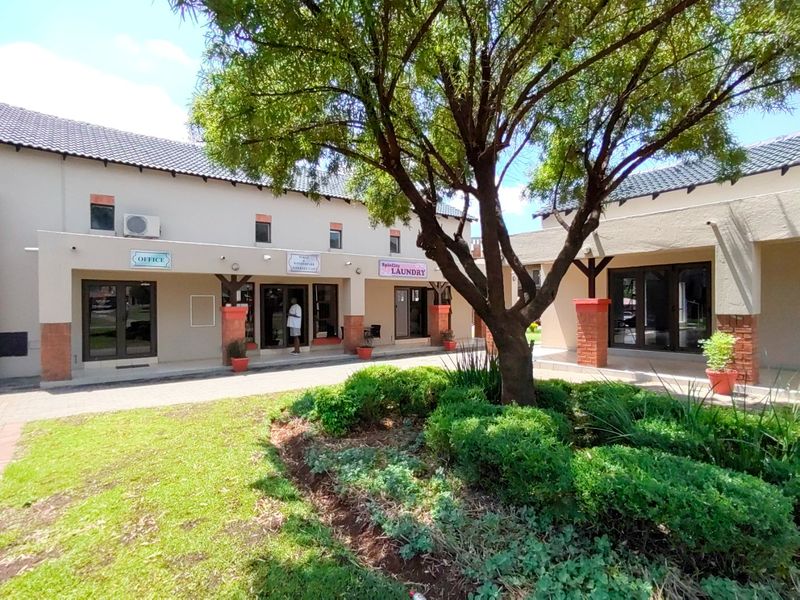 A well-maintained exterior courtyard area featuring paved walkways, landscaped garden beds, and multiple commercial-style glass entrance doors under a tiled roof.