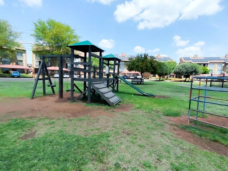 Communal outdoor playground area featuring a wooden climbing frame, slide, and swing set situated within the apartment complex grounds.