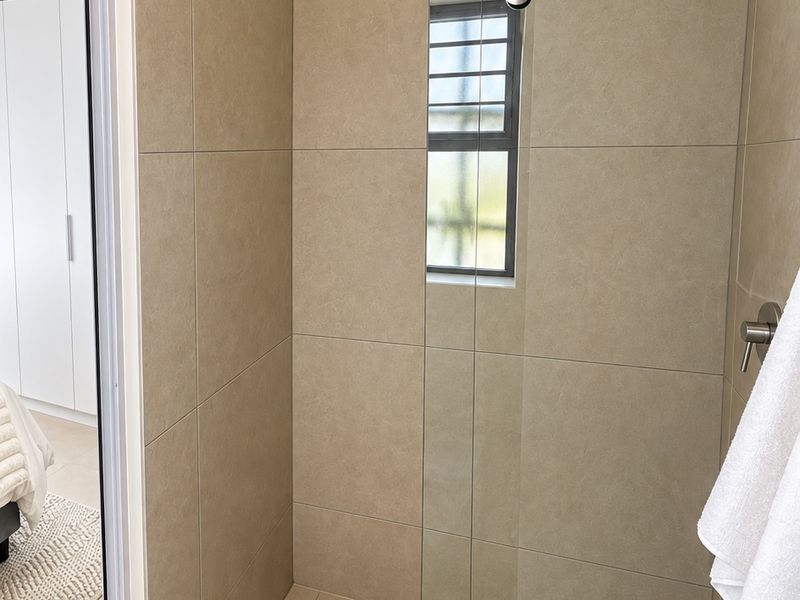 A modern, well-maintained walk-in shower featuring neutral large-format wall tiles, a glass screen, and a window for natural light.