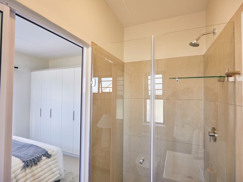 A modern bathroom featuring a glass-enclosed shower with neutral tiling, adjacent to a bedroom with built-in wardrobe storage.