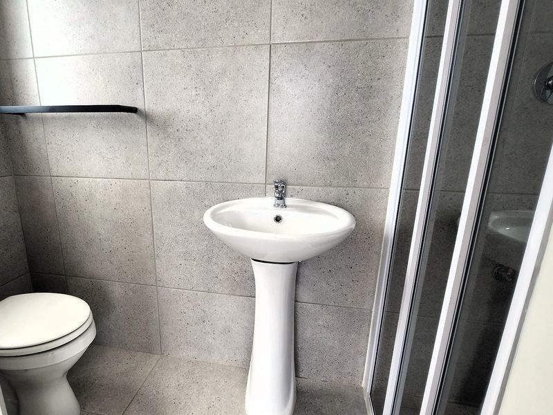 A modern, clean bathroom featuring a pedestal basin, toilet, and a glass-enclosed shower with neutral grey wall and floor tiles.
