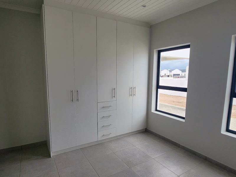 A bright, modern bedroom featuring neutral tiled flooring, clean white built-in cupboards, and large aluminium-framed windows.
