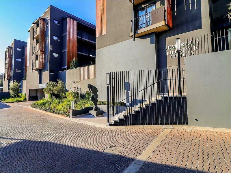 Modern apartment complex exterior featuring contemporary facade design, paved driveway, secure metal fencing, and landscaped garden areas leading to the lifestyle centre entrance.