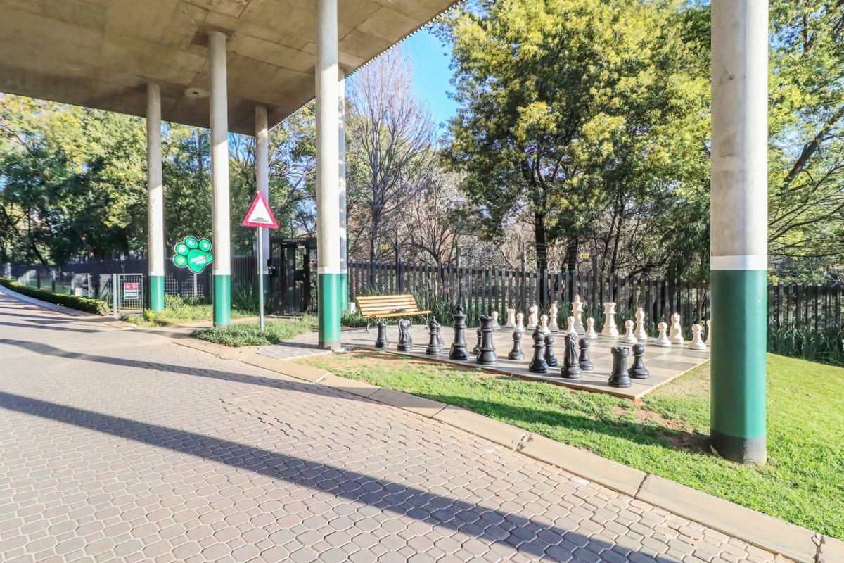 A well-maintained outdoor communal area featuring a paved driveway, concrete structural pillars, and a landscaped garden with a large outdoor chess set.
