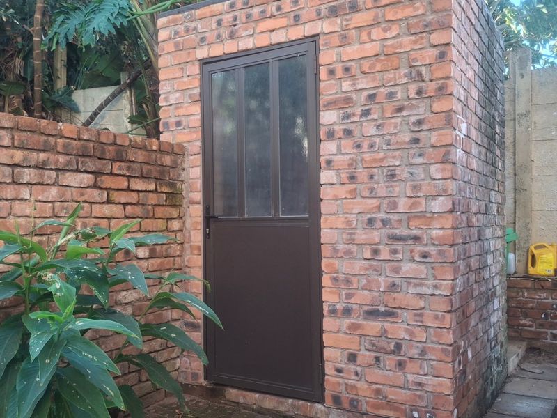 A small, standalone utility structure constructed from exposed red brick with a metal-framed door, situated in an outdoor garden area.