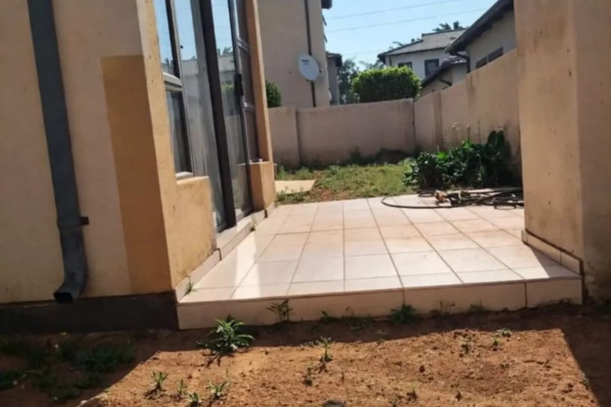 Rear exterior view of the apartment featuring a tiled patio area, sliding glass doors, and an unlandscaped garden space.