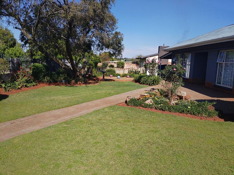 A well-maintained rear garden featuring a paved walkway, established trees, and landscaped flower beds adjacent to the house exterior.