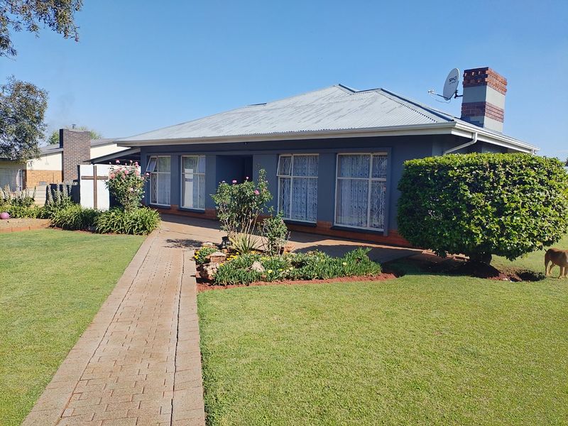 A well-maintained single-story home featuring a paved walkway, landscaped front garden, and a prominent brick chimney under a corrugated metal roof.