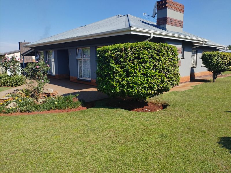 A well-maintained single-story house featuring a paved front patio, brick chimney, and established garden with manicured shrubs and lawn.