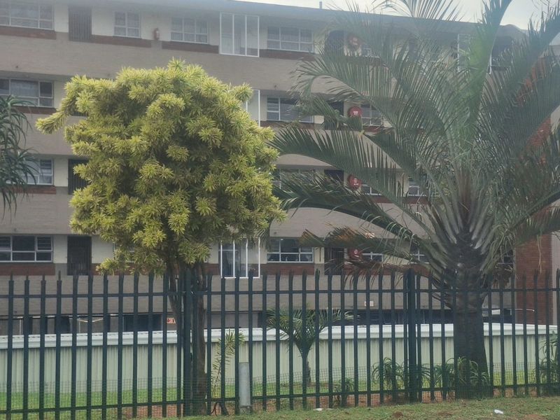 Exterior view of a multi-story apartment building featuring a secure metal perimeter fence and established landscaping.