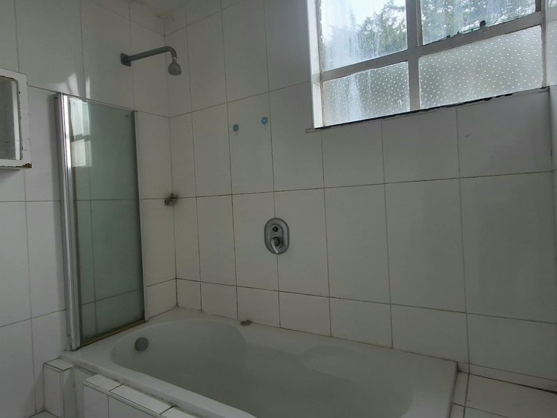 A dated bathroom featuring a built-in bathtub with a shower mixer, glass screen, and wall-mounted storage cabinet, requiring cosmetic updates to modernize the space.